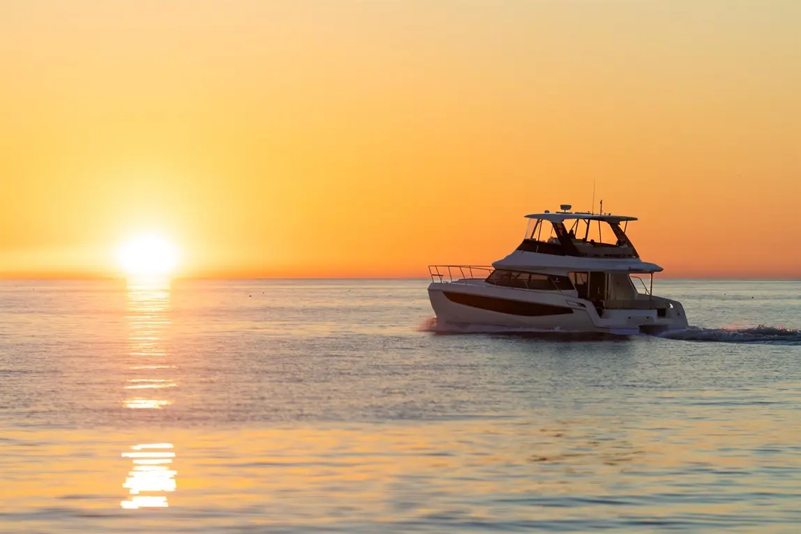 A boat glides across calm waters at sunset, reflecting golden hues from the sun on the horizon.